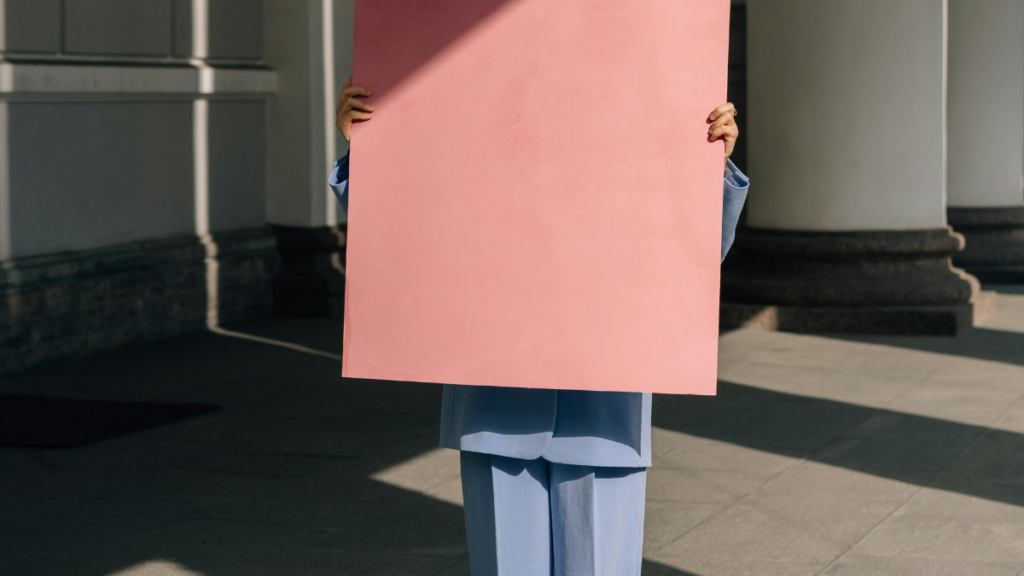 Eine Frau im Anzug, die vor ihrem Gesicht und Oberkörper ein leeres, rosafarbenes Plakat hält.